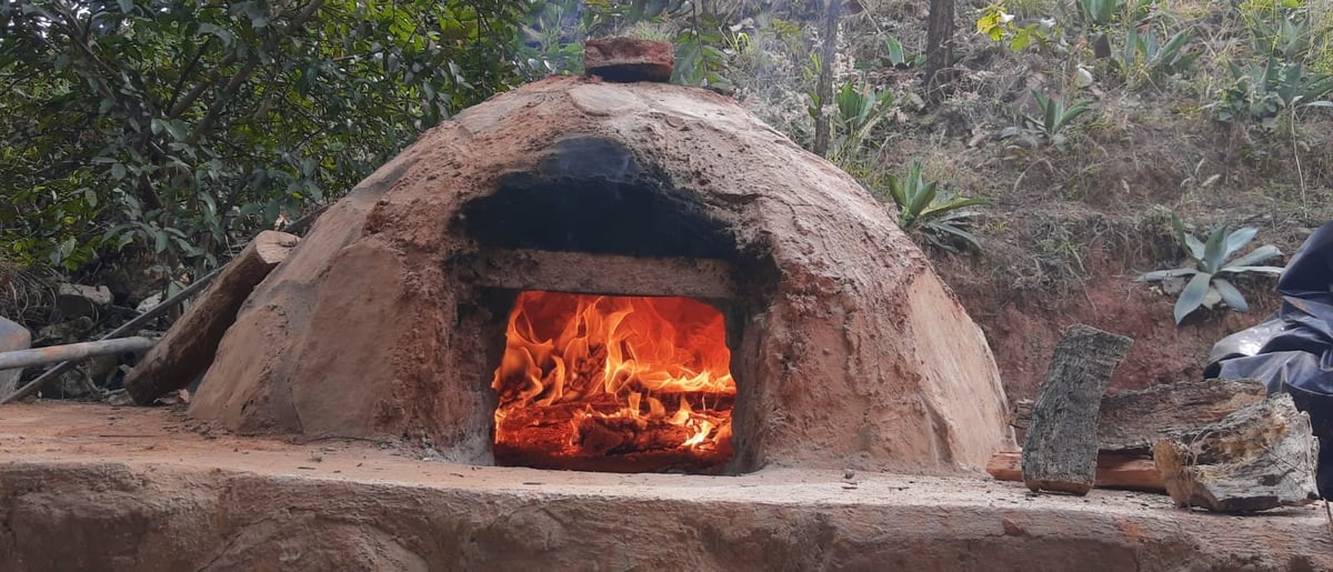 Clay dome oven with bright orange flames burning inside, surrounded by dry vegetation and wood storage